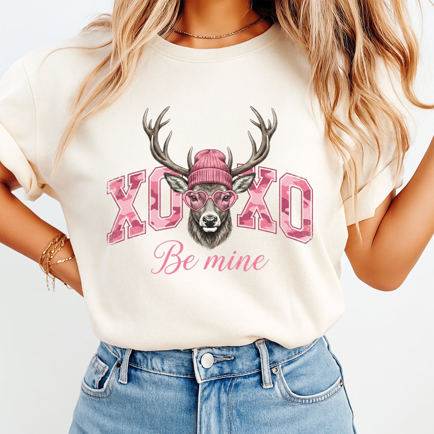 Cream shirt mockup showing Valentine’s Day deer graphic with XOXO lettering, pink beanie, heart sunglasses, and Be Mine text