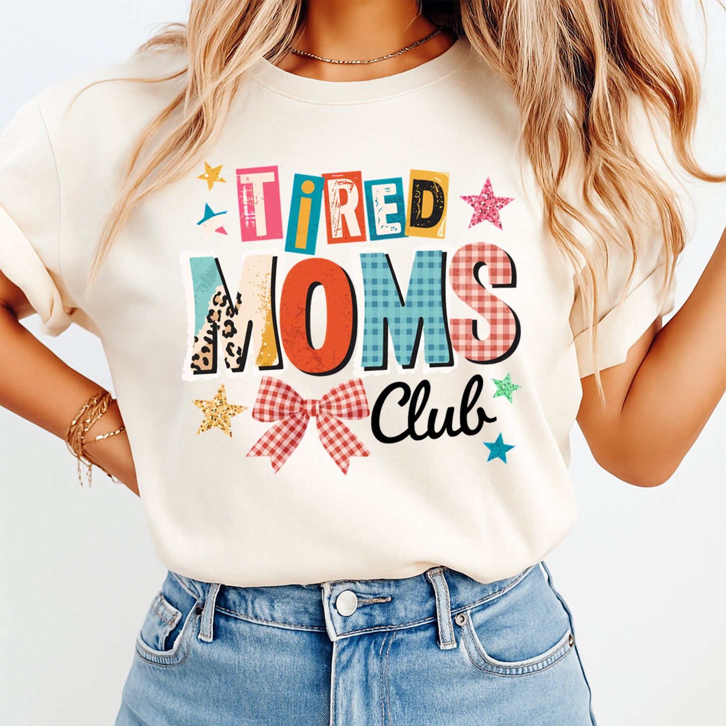 Woman wearing a cream shirt with tired moms club ready to press dtf graphic featuring colorful mixed pattern letters bow and stars