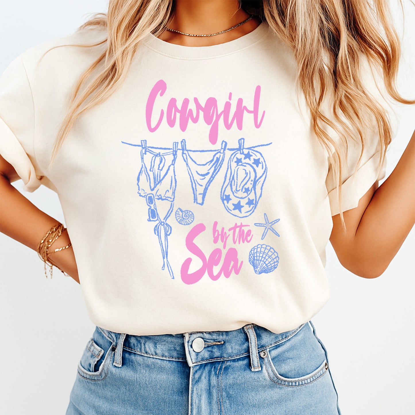 Person wearing a t-shirt with 'Cowgirl by the Sea' design and denim shorts on a light background