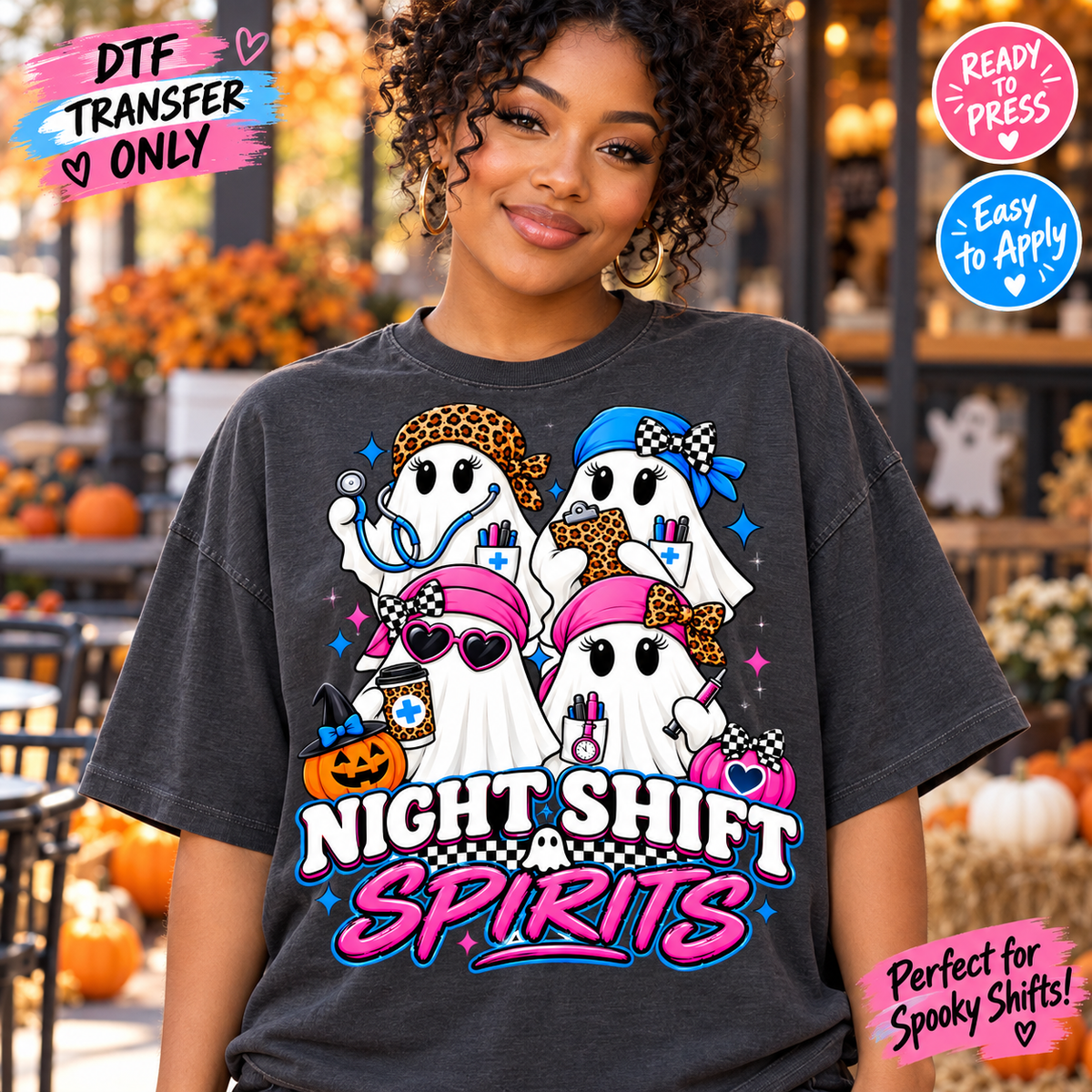 A woman wearing a dark charcoal gray graphic T-shirt with a colorful Halloween night shift design that reads 'Night Shift Spirits' with cartoon ghosts, pumpkins, and stars. The background is a blurred autumn storefront with pumpkins and decorations.