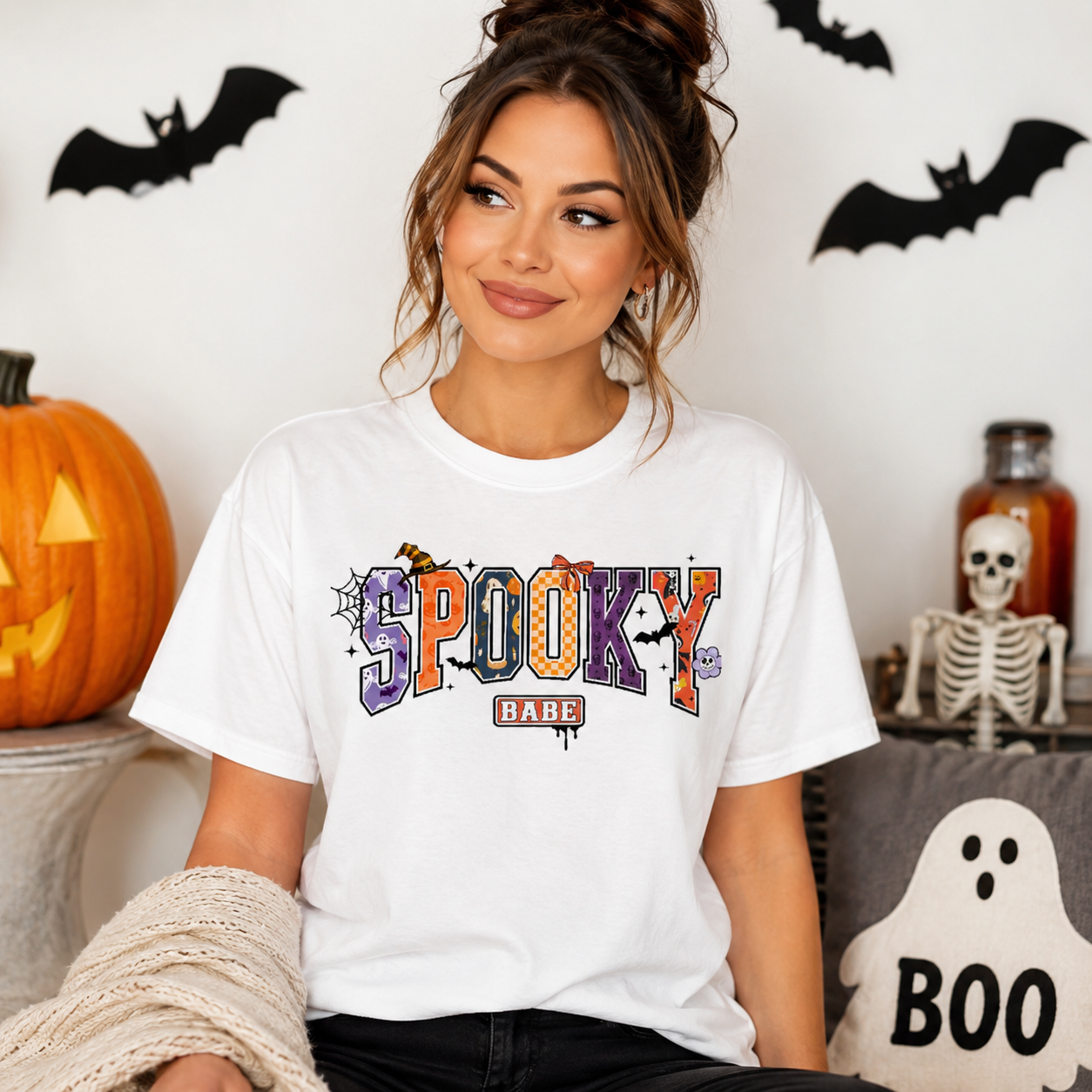 A woman wearing a white graphic t-shirt with 'Spooky Bare' printed in colorful letters, posed in a Halloween-themed room with pumpkins, bats, and a skeleton.