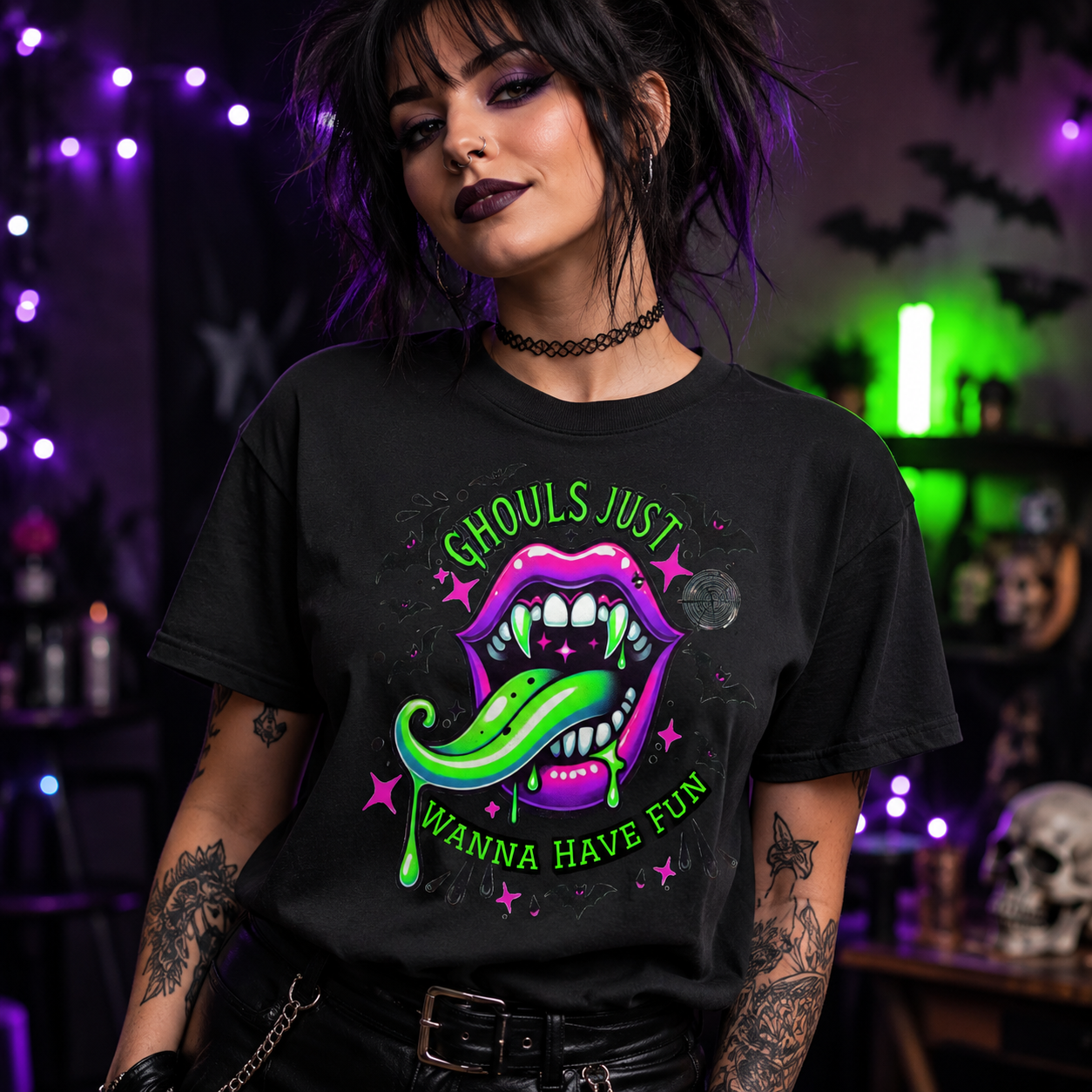 A woman wearing a black graphic t-shirt with a colorful design of vampire lips, a green tongue, and the text 'GHOULS JUST WANNA HAVE FUN'. The background is a dark, spooky room with purple and green lights, skulls, and bats.