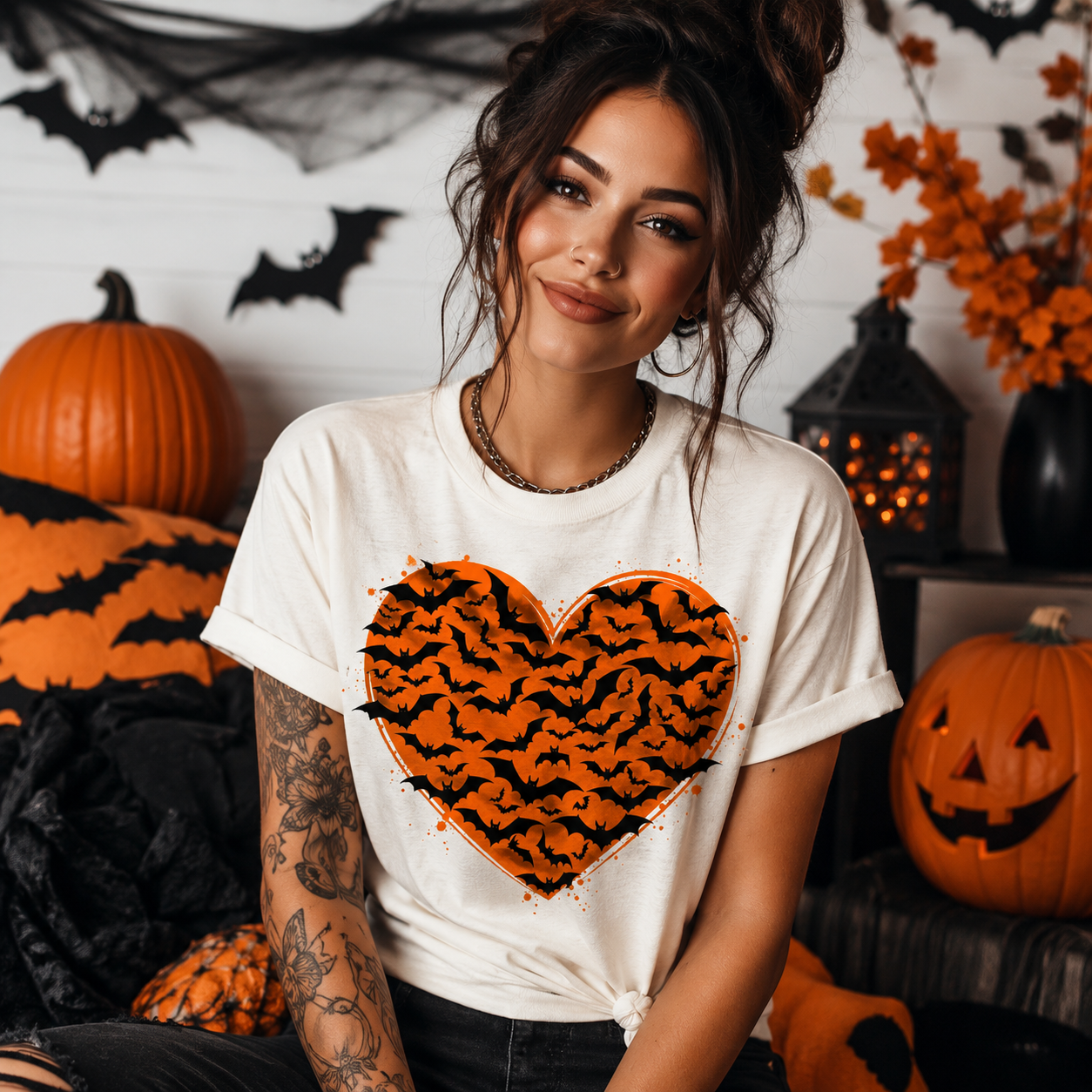 A woman wearing a white t-shirt with a large orange and black patterned heart graphic on the front, posed in a Halloween-themed setting with pumpkins, bats, and a lantern.