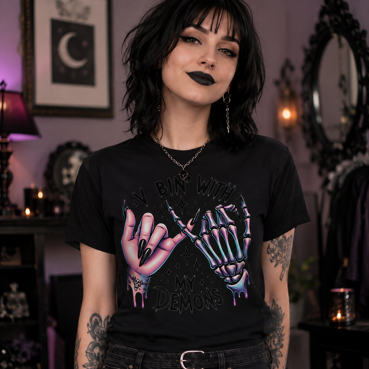 A woman with short black hair and gothic makeup wearing a black graphic t-shirt that reads 'MY BIN WITH MY DEMONS' and features two skeletal hands making a shaka sign. She is standing in a dimly lit, gothic-themed room with candles, mirrors, and framed art.