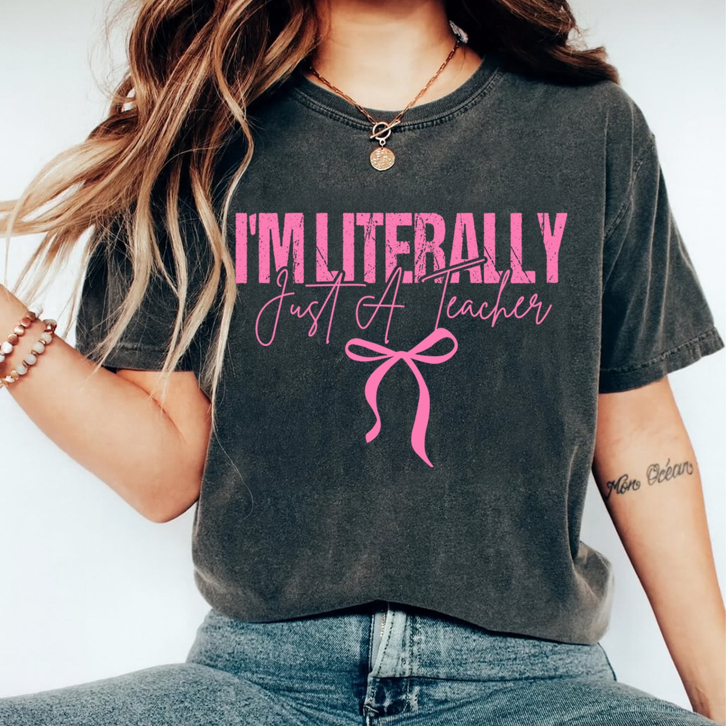 Dark gray t shirt mockup showing the text I’m Literally Just A Teacher in pink lettering with bow detail, teacher graphic tee