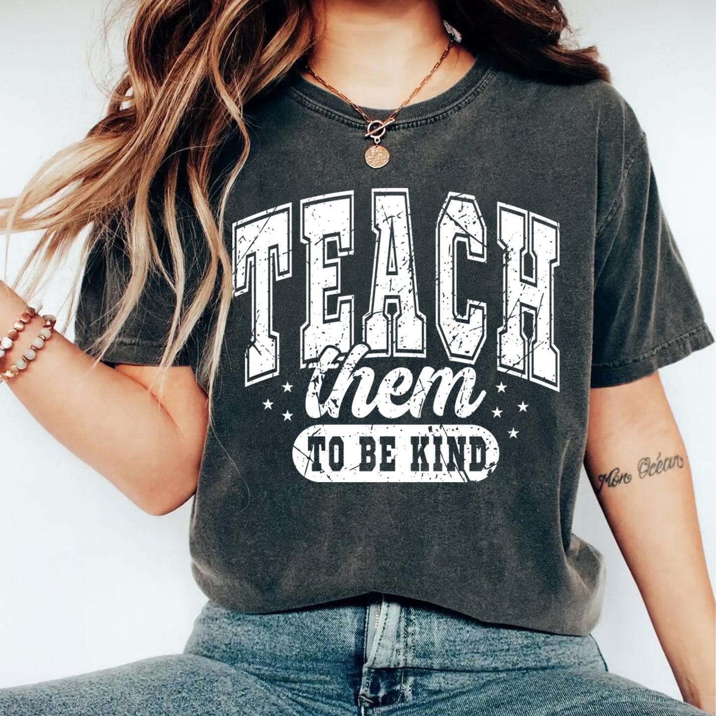 Teach them to be kind teacher graphic displayed on a dark charcoal t shirt mockup with bold white lettering and vintage style