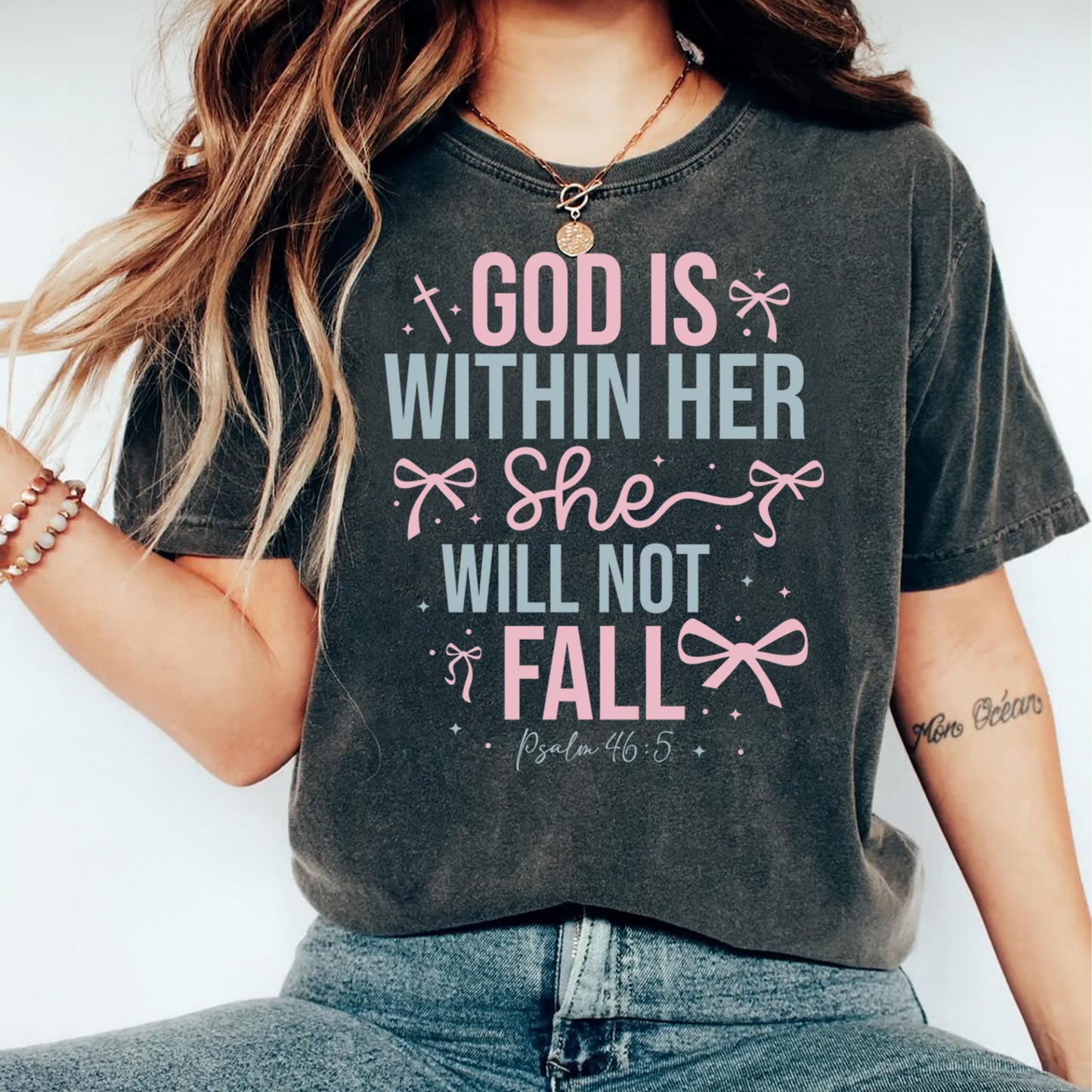 Christian faith shirt mockup showing God Is Within Her She Will Not Fall scripture design with feminine bows and pastel lettering on dark tee