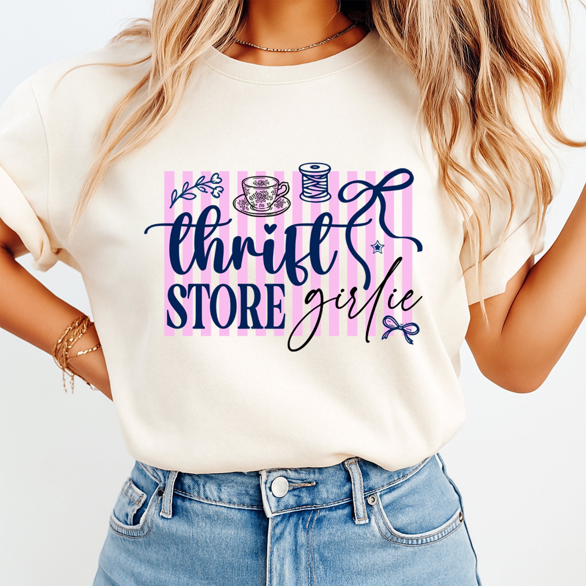 Woman wearing a cream shirt with thrift store girlie graphic featuring pink stripes, tea cup, thread spool, bows, and floral accents, ready to press dtf mockup