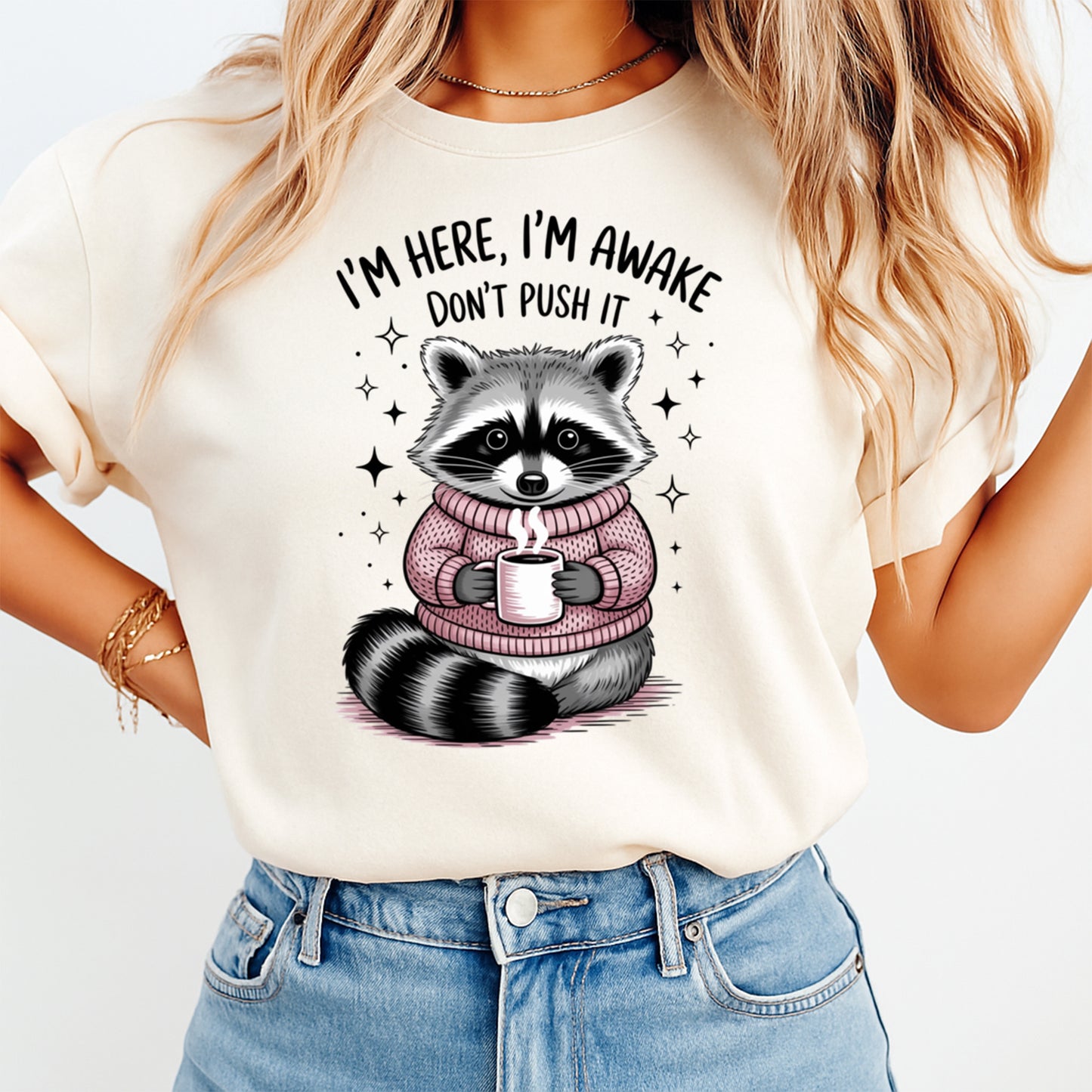 Mockup shirt featuring a cute raccoon in a pink sweater holding a coffee mug with the phrase I’m here I’m awake don’t push it