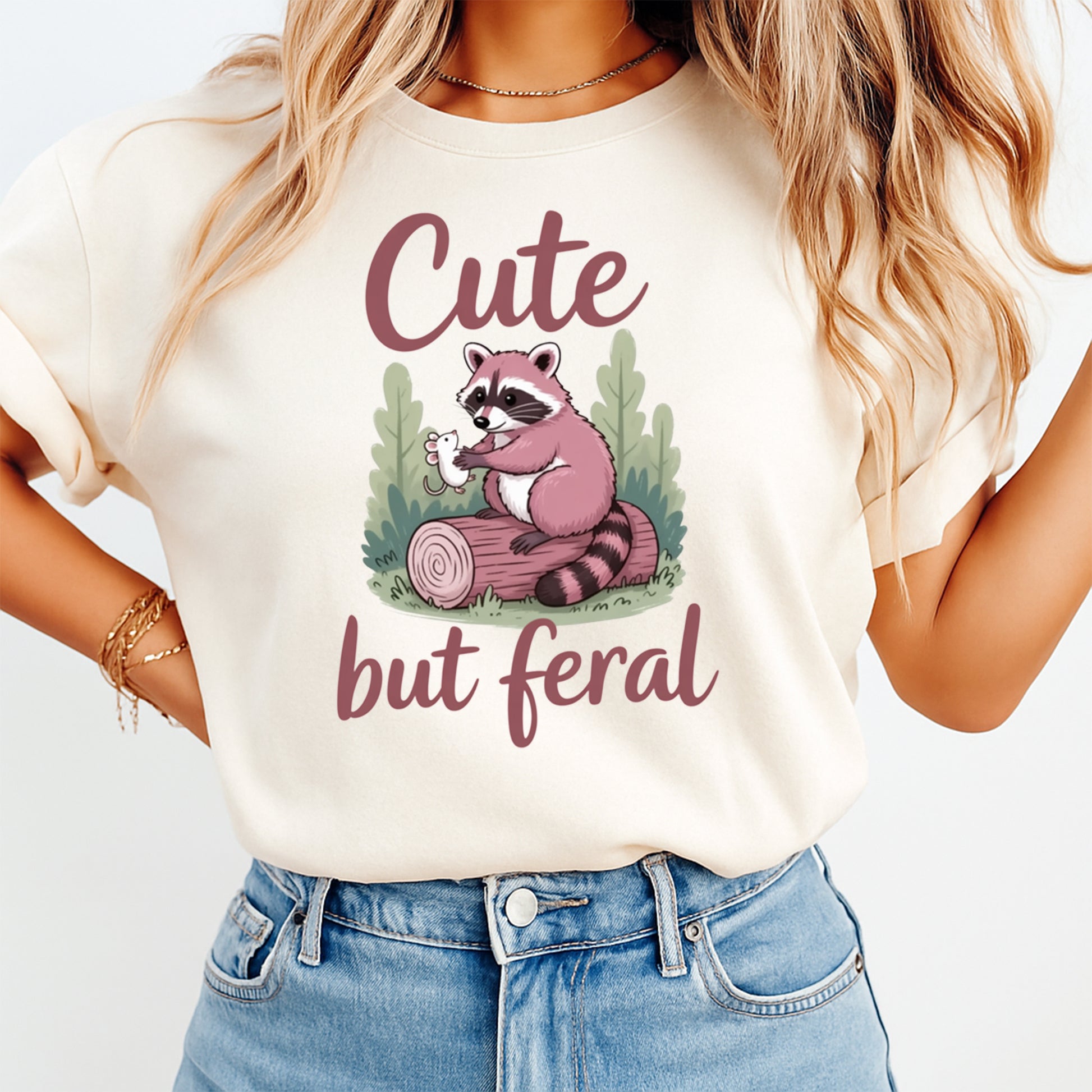 Mockup shirt featuring a pink raccoon sitting on a log holding a tiny mouse with the phrase Cute but feral