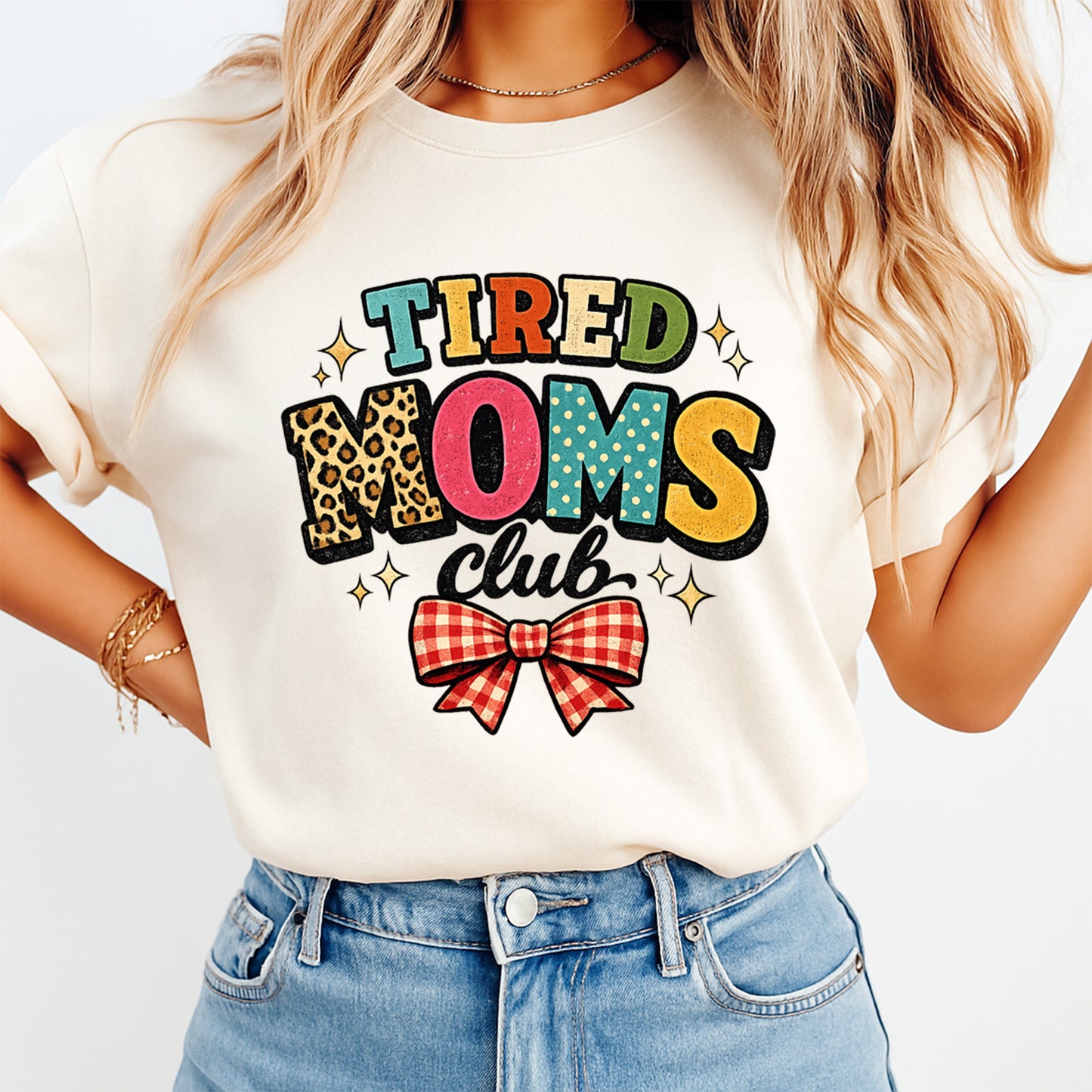 Mockup image of a cream shirt featuring the phrase Tired Moms Club in colorful retro letters with leopard print, sparkles, and a red gingham bow