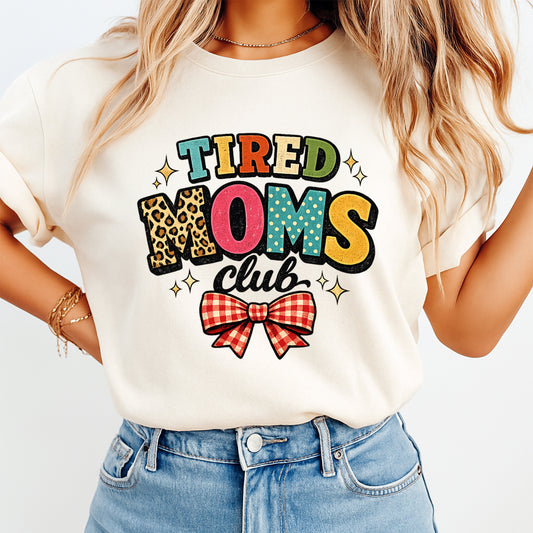Mockup image of a cream shirt featuring the phrase Tired Moms Club in colorful retro letters with leopard print, sparkles, and a red gingham bow
