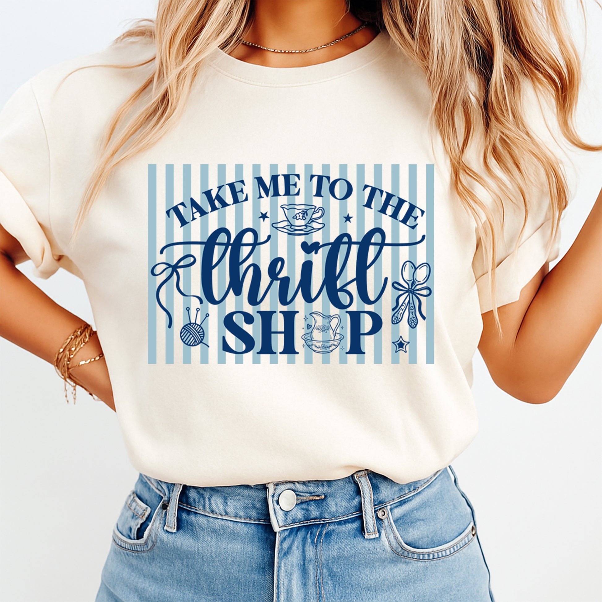 Mockup of a cream shirt featuring a thrift shop ready to press dtf transfer with blue vintage lettering, striped background, teacups, yarn ball, bow, and the phrase Take Me To The Thrift Shop.