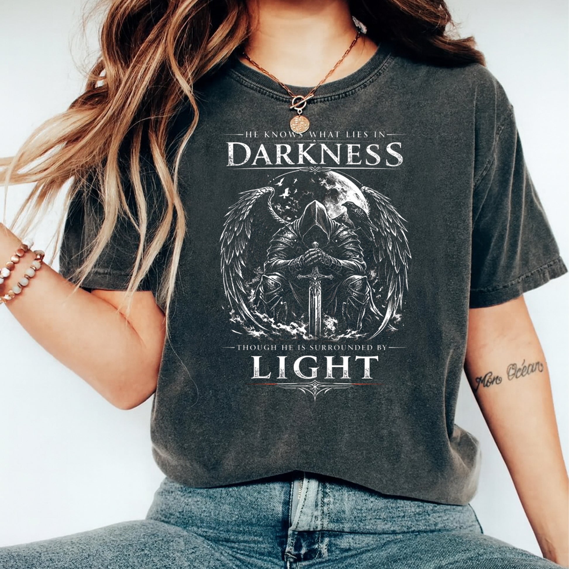 Mockup of a dark shirt featuring an inspirational warrior ready to press dtf transfer with angel wings, moon, sword, and the phrase He knows what lies in darkness though he is surrounded by light.