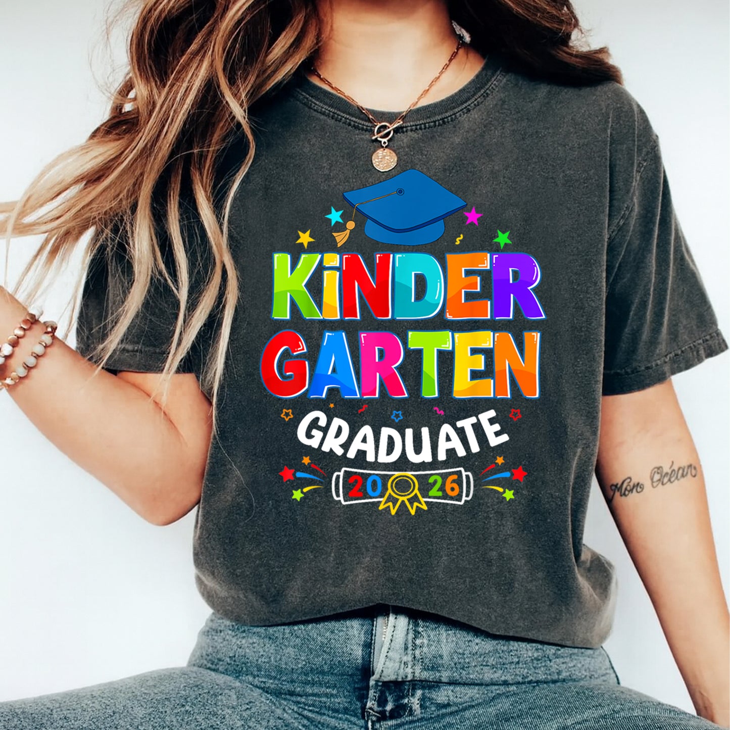 Mockup of black shirt featuring Kindergarten Graduate 2026 ready to press dtf transfer with colorful lettering, blue graduation cap, stars, and school celebration artwork.