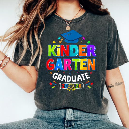 Mockup of black shirt featuring Kindergarten Graduate 2026 ready to press dtf transfer with colorful lettering, blue graduation cap, stars, and school celebration artwork.