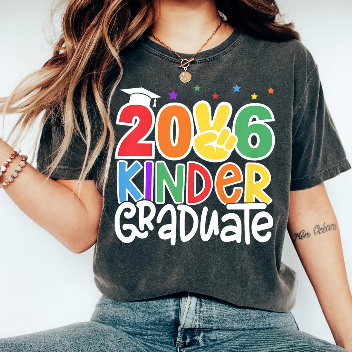 Mockup of black shirt featuring 2026 Kinder Graduate ready to press dtf transfer with colorful rainbow lettering, stars, cap, and peace sign artwork.