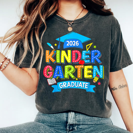 Black shirt mockup featuring a Kinder Garten Graduate 2026 ready to press dtf transfer with colorful letters, graduation cap, school supplies, and blue banner.