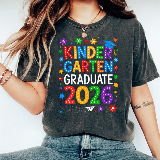 Black shirt mockup featuring a Kinder Garten Graduate 2026 ready to press dtf transfer with rainbow letters, graduation cap, stars, and flower accents.
