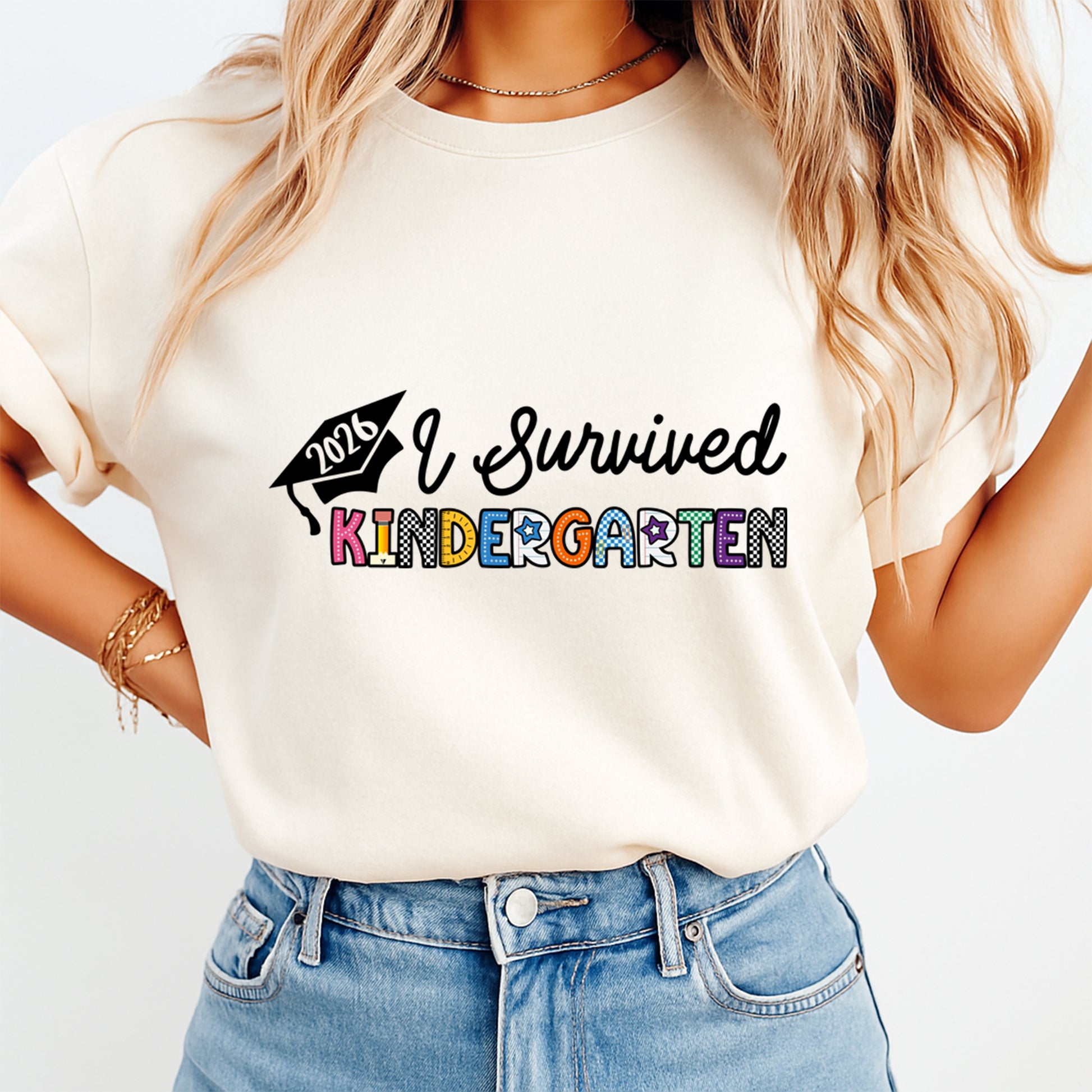 Cream shirt mockup featuring an I Survived Kindergarten 2026 ready to press dtf transfer with graduation cap and colorful school lettering.