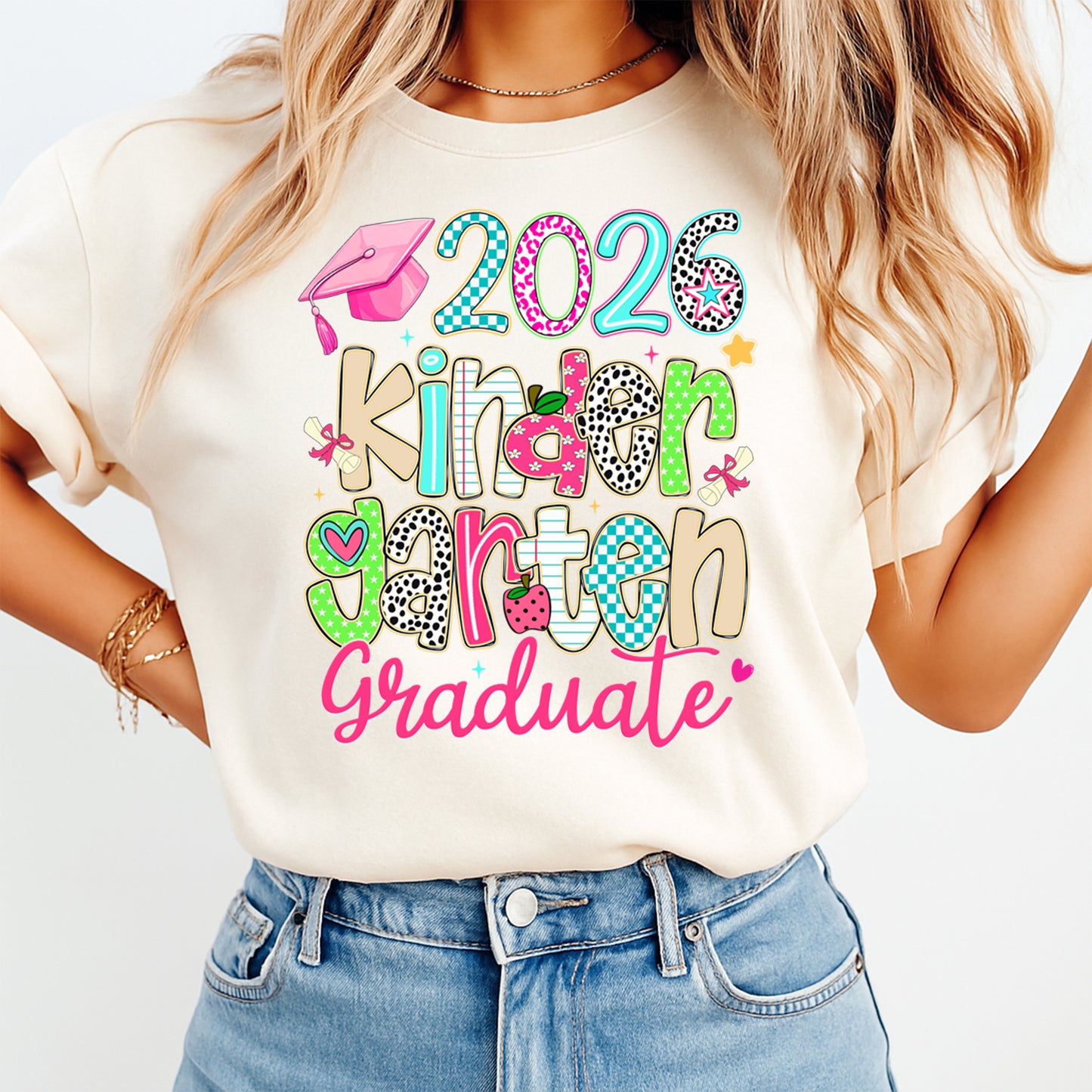Mockup of a cream shirt featuring a kindergarten graduation ready to press dtf transfer with the phrase 2026 kinder garten Graduate, colorful letters, pink cap, diplomas, apple, and stars.