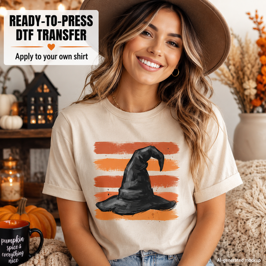 A woman wearing a beige t-shirt with a Halloween graphic of a black witch hat on orange striped brushstrokes, styled with a brown hat in a cozy fall-themed room setting.