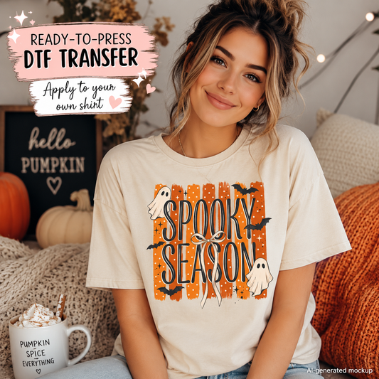 A woman wearing a cream-colored t-shirt with a 'Spooky Season' graphic print featuring orange pumpkins, black bats, and white ghosts. The image is a mockup with promotional text overlays on the top left and a cozy home background with pumpkins and a mug.