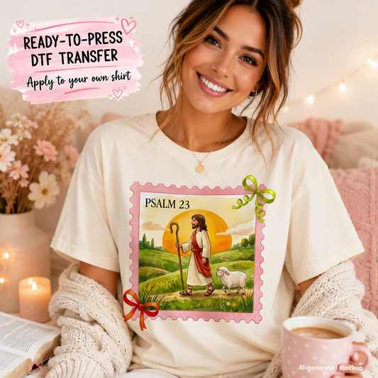 A smiling woman wearing a cream-colored t-shirt with a colorful graphic of Jesus as a shepherd and a white sheep under a sunset, framed by a pink border with 'PSALM 23' text. She is holding a mug in a cozy, warmly lit bedroom setting with candles and flowers.