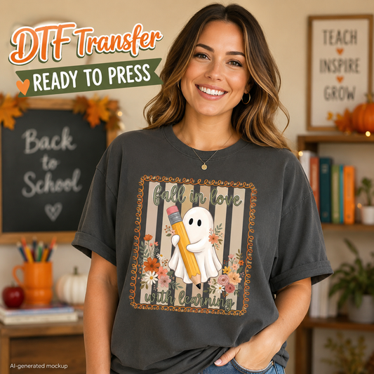 A woman wearing a dark charcoal gray t-shirt with a 'Fall in love, write happily' ghost graphic holding a yellow pencil. The image is a digital mockup with 'DTF Transfer Ready to Press' text overlay in the top left, set against a cozy, warm autumn-themed classroom background.