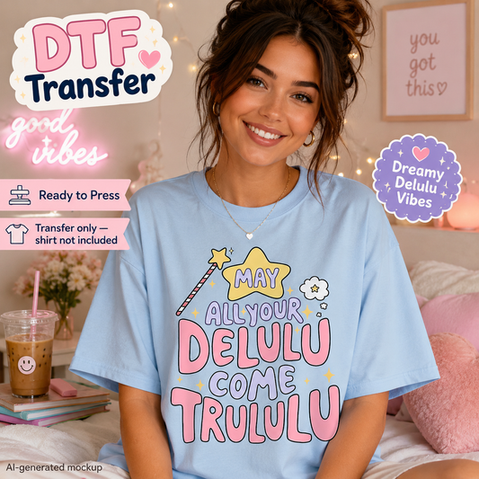 A woman wearing a light blue graphic T-shirt with a colorful 'May All Your Delulu Come Trululu' design, posed in a cozy bedroom setting with decorative text overlays.