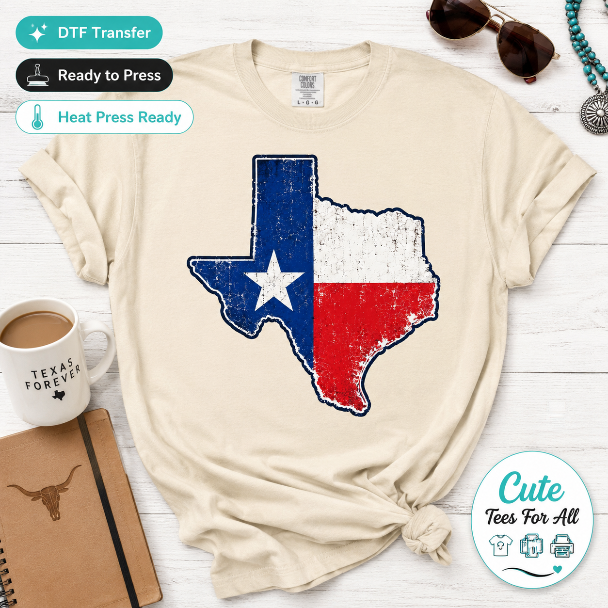 A beige short-sleeve t-shirt with a distressed Texas flag graphic, styled in a flat-lay arrangement on a white wood background with coffee, sunglasses, a notebook, and a decorative badge.