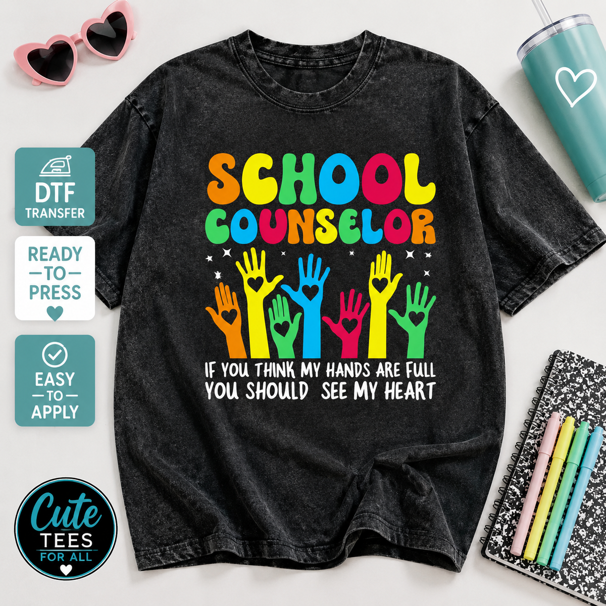 A black acid-wash graphic t-shirt featuring a colorful 'SCHOOL COUNSELOR' design with raised hands and the text 'IF YOU THINK MY HANDS ARE FULL YOU SHOULD SEE MY HEART'. The t-shirt is displayed in a styled flat-lay arrangement with props including pink heart-shaped sunglasses, a teal tumbler, a notebook with colored pens, and promotional badges on the left side.