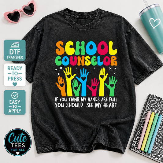 A black acid-wash graphic t-shirt featuring a colorful 'SCHOOL COUNSELOR' design with raised hands and the text 'IF YOU THINK MY HANDS ARE FULL YOU SHOULD SEE MY HEART'. The t-shirt is displayed in a styled flat-lay arrangement with props including pink heart-shaped sunglasses, a teal tumbler, a notebook with colored pens, and promotional badges on the left side.