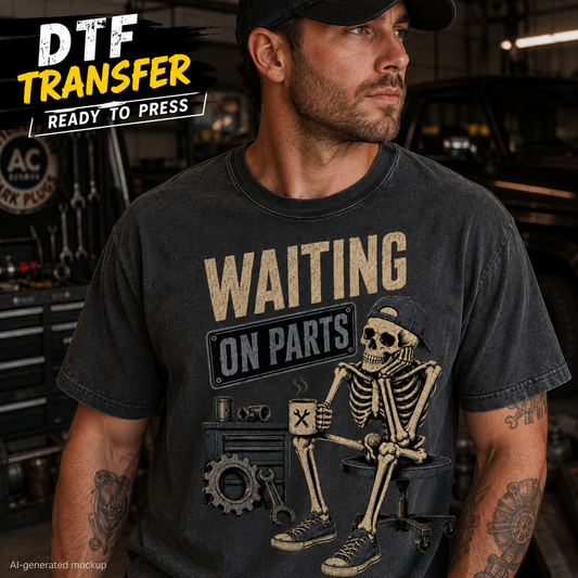 A man wearing a black graphic T-shirt with a distressed print that reads 'WAITING ON PARTS' above a skeleton mechanic sitting at a desk with tools. The background is a garage or workshop setting with tools and vehicles.