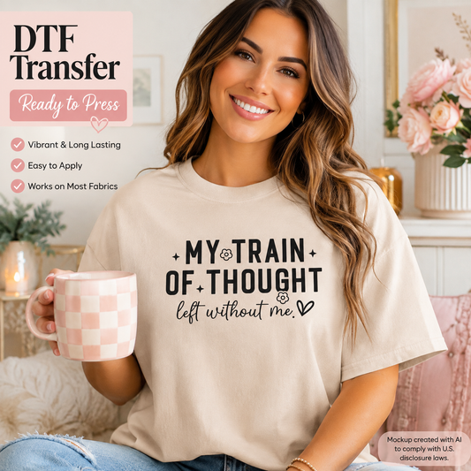 A woman wearing a cream-colored t-shirt with a black graphic print that reads 'MY TRAIN OF THOUGHT left without me' and holding a pink and white checkered mug. The image features promotional text overlays for an 'DTF Transfer' product on a cozy, decorated background.