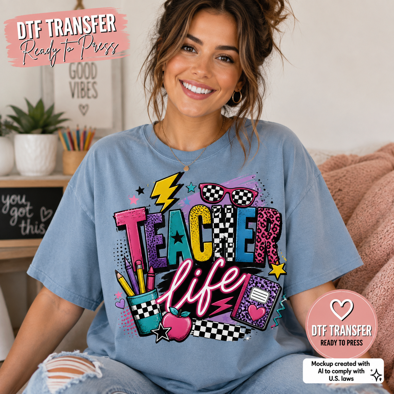 A woman wearing a light blue graphic T-shirt with a colorful 'Teacher Life' design, seated in a cozy indoor setting. The graphic features playful text, stars, checkered patterns, and school-themed illustrations in multiple bright colors.