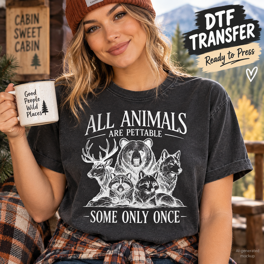 A woman wearing a dark charcoal gray graphic t-shirt with a white distressed print of various animals and the text 'ALL ANIMALS ARE PETTABLE SOME ONLY ONCE'. She is holding a white mug that says 'Good People Wild Places' and wearing a brown beanie.