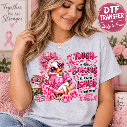 A woman wearing a light heather gray t-shirt with a colorful graphic of a pink breast cancer awareness duck and text, posed in a cozy room setting with a 'DTF TRANSFER Ready to Press' badge overlay.
