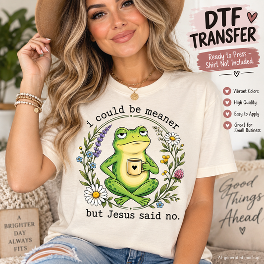Woman wearing a cream shirt featuring a funny green frog holding a coffee mug surrounded by flowers with the phrase I Could Be Meaner But Jesus Said No as a DTF transfer mockup.