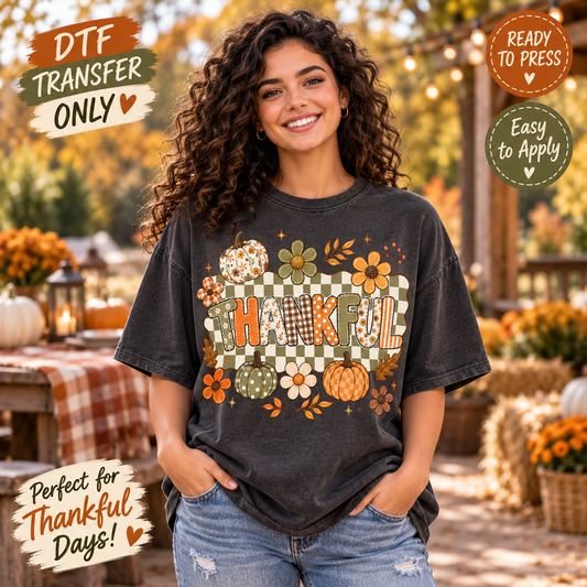 A woman wearing a dark charcoal gray graphic t-shirt with a fall-themed 'HAFUL' design featuring pumpkins, flowers, and a checkered pattern. The background is a warm, autumnal outdoor setting with tables, pumpkins, and string lights.