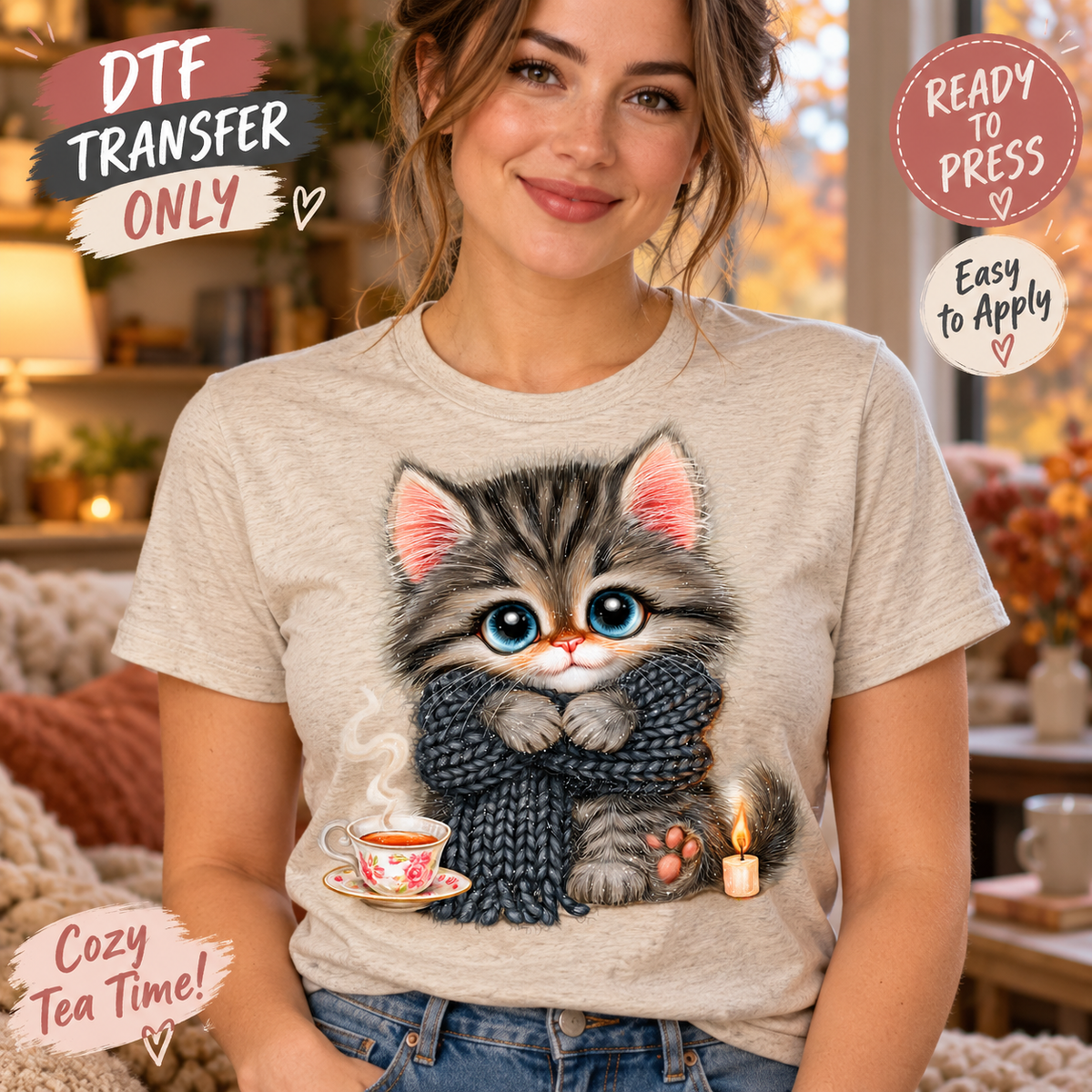 A woman wearing a light beige graphic t-shirt featuring a cute cartoon kitten wrapped in a dark gray knit sweater, holding a teacup and next to a candle. The background is a cozy, warm-toned living room.