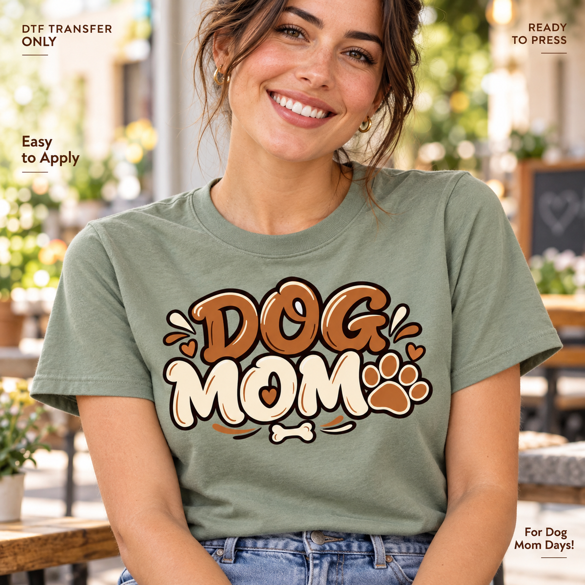 Lifestyle mockup of a woman wearing a sage green shirt showing Dog Mom in brown and cream lettering with paw print, hearts, and a bone. DTF transfer only; shirt not included.
