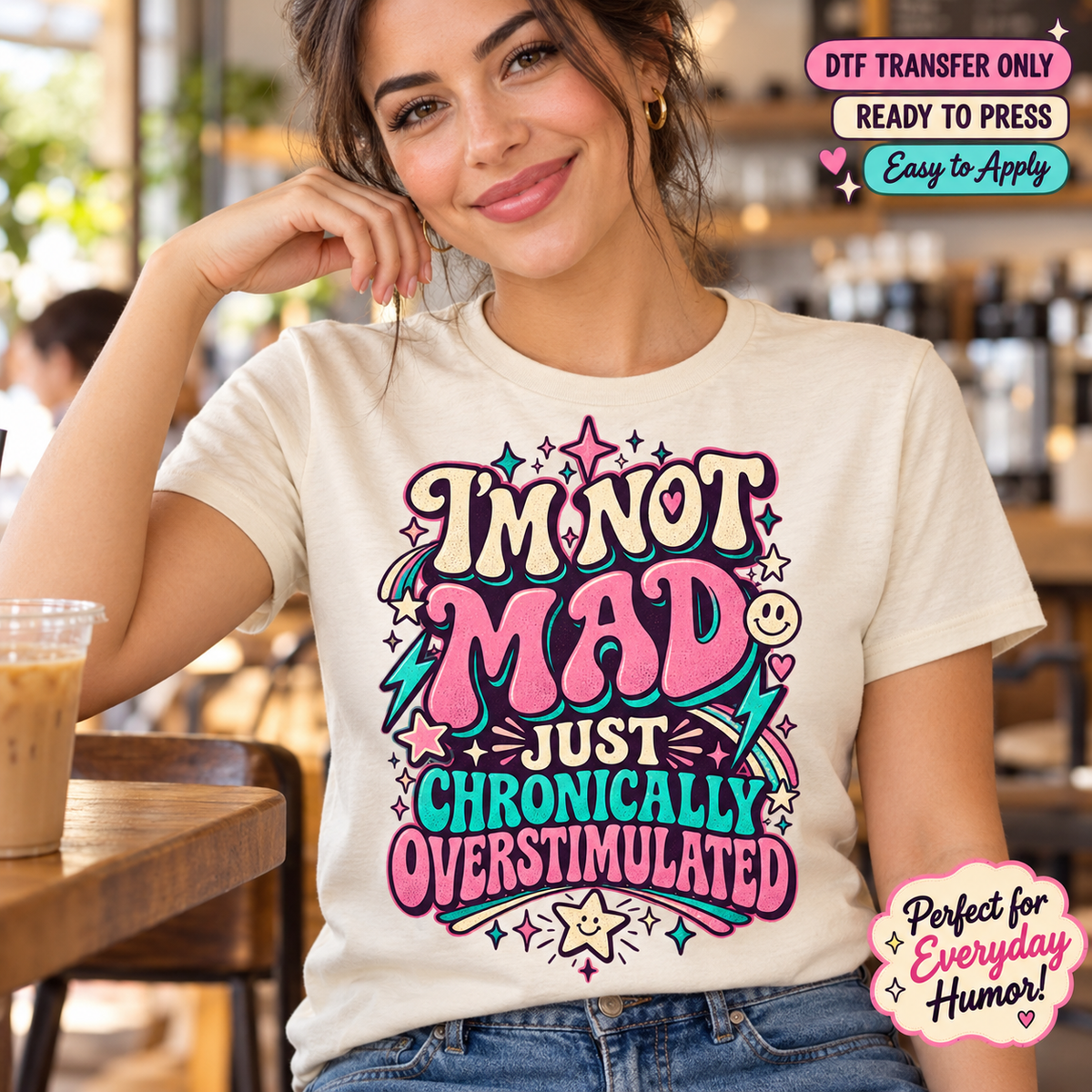Lifestyle mockup of a woman wearing a cream shirt showing I'm Not Mad Just Chronically Overstimulated lettering with pink and teal accents, stars, and lightning bolts. DTF transfer only; shirt not included.