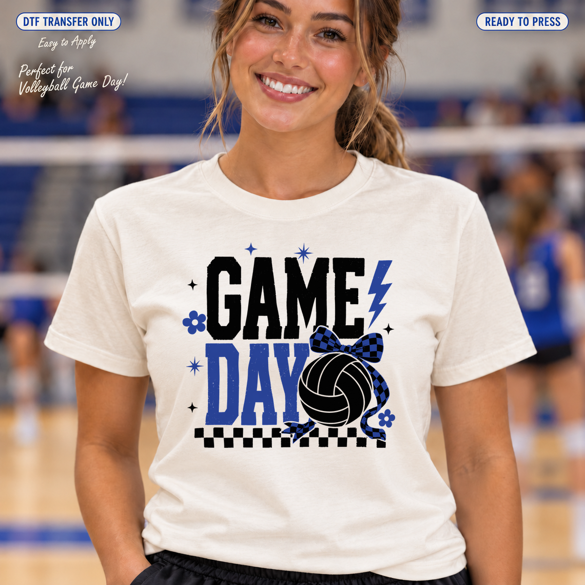 Woman in a cream shirt with black Game and blue Day lettering, a black volleyball, blue bow and checkerboard accent in a gym.