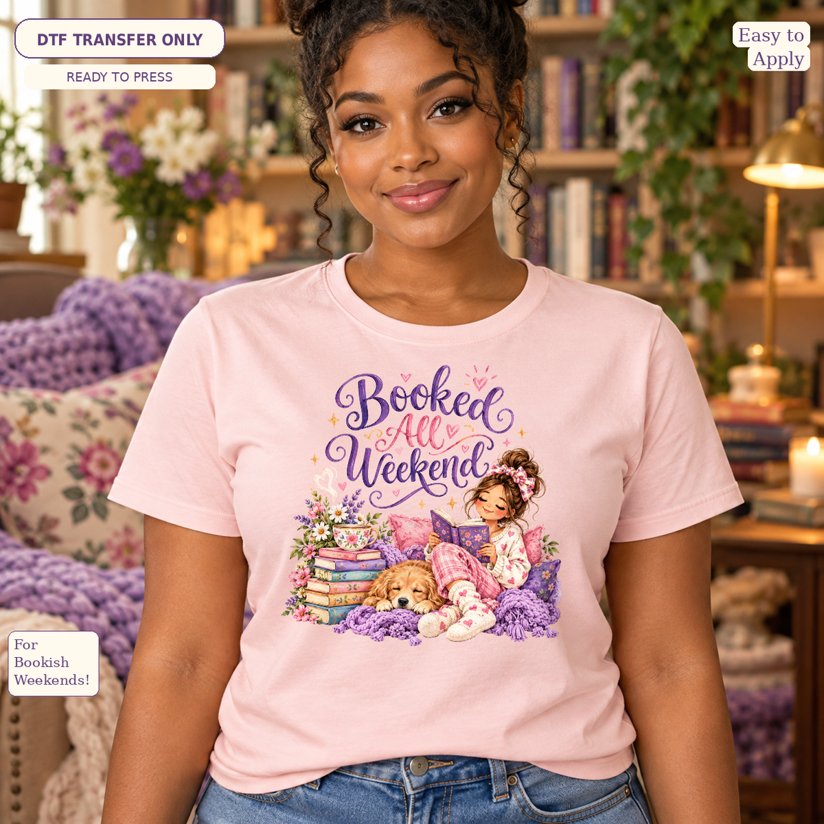 Woman wearing a pink tee with purple Booked All Weekend lettering and a cozy reader beside a stack of books.