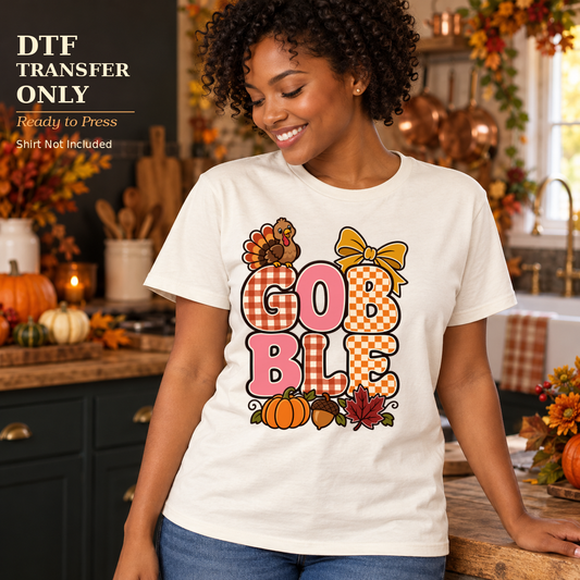 Woman wearing a white shirt with patchwork Gobble lettering, a small turkey, yellow bow, and pumpkins in a fall kitchen.