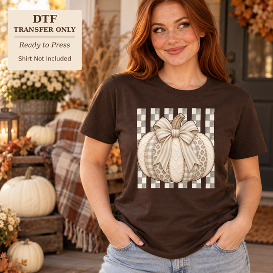 Woman wearing a brown shirt with a neutral patterned pumpkin, large cream bow, and gingham background on a porch.