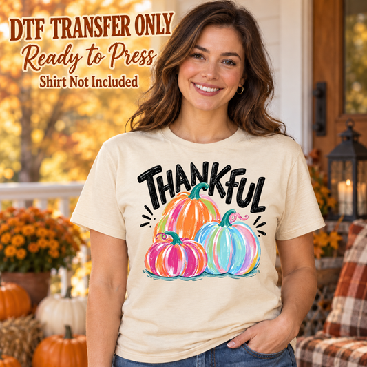 Woman in an ivory tee with black Thankful lettering above three brightly colored pink, orange and blue pumpkins, labeled transfer only.