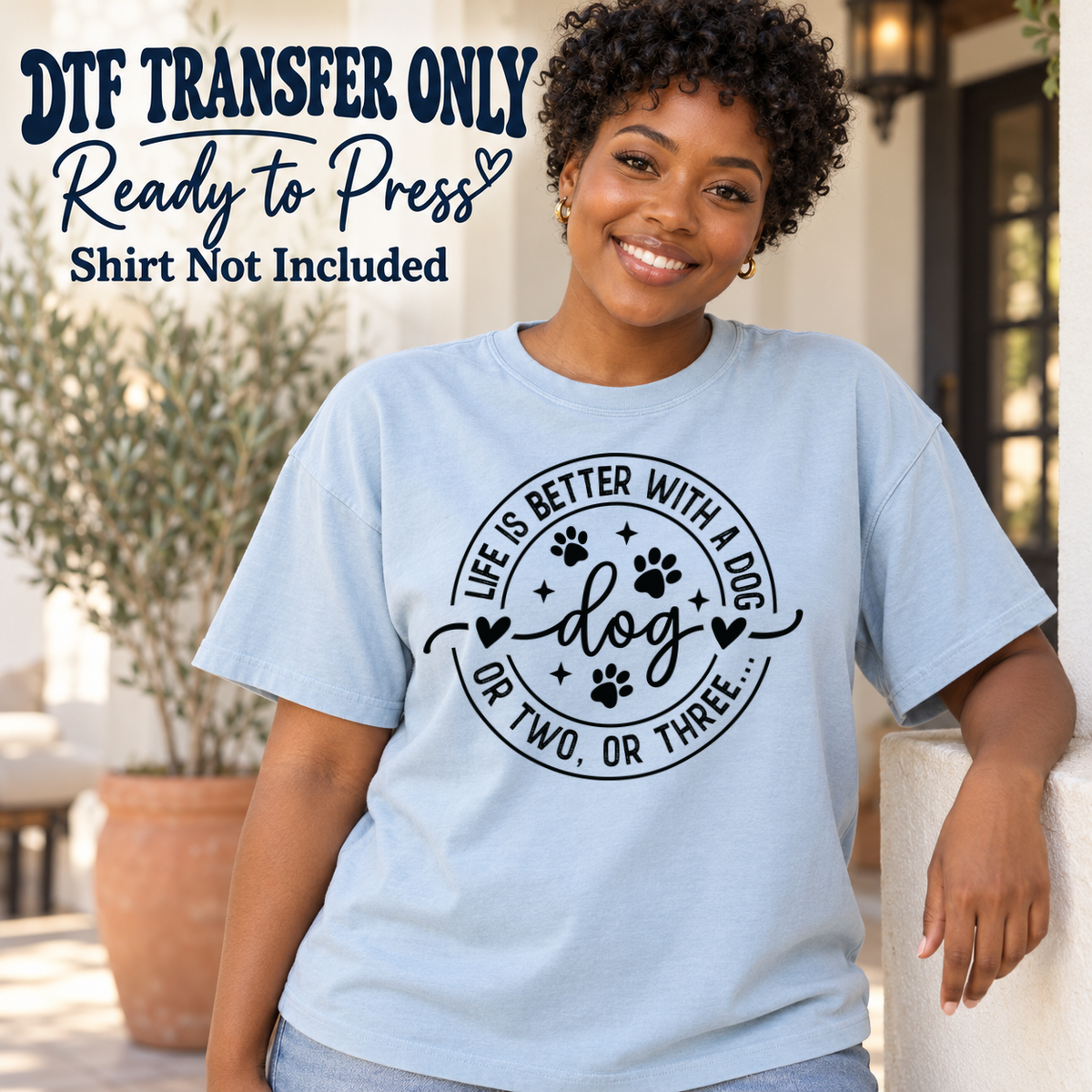 Black circular Life's Better With A Dog Or Two Or Three graphic with paw prints, shown on a light blue shirt model mockup; physical DTF transfer only, shirt not included.