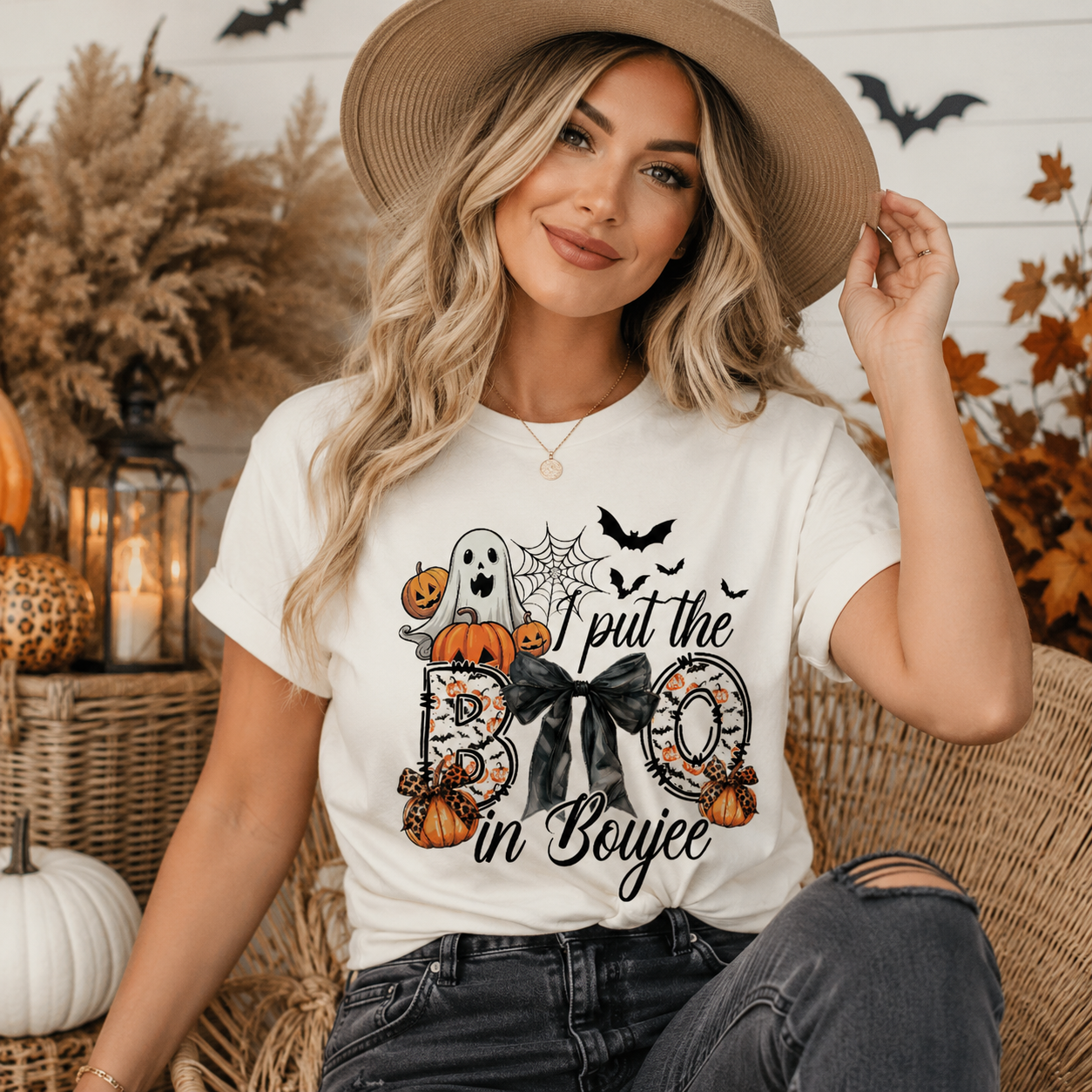 A woman wearing a white graphic t-shirt with a Halloween design that reads 'I put the BOYjee in Boyjee', styled with a wide-brimmed straw hat and dark jeans in a cozy, autumnal setting.